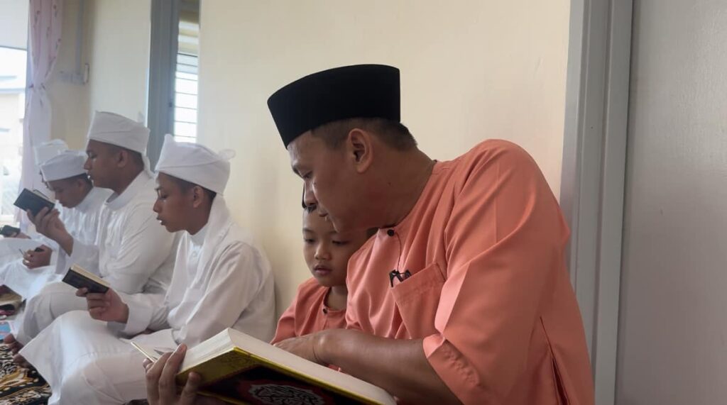 Ayah memakai baju oren lembut menunjukkan anak membaca Al-Baqarah penuh bersama pelajar tahfiz sempena pembukaan Taska Kasih Fitri (TKF) Membakut.